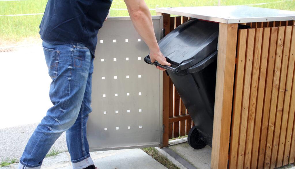 Mann in Jeans und schwarzem T-Shirt stellt eine schwarze Mülltonne in eine stilvolle Mülltonnenbox aus Holz und Metall auf einem Gehweg.