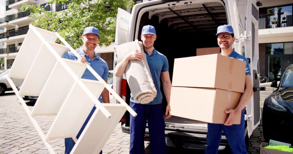 Drei Möbelpacker in blauen Uniformen stehen lächelnd vor einem Lieferwagen. Sie tragen Umzugskartons, einen Teppich und ein Regal, bereit für den Möbeltransport.